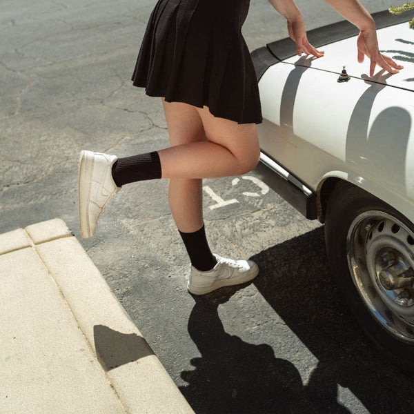 Dame stützt sich auf ein weißes Auto, trägt einen schwarzen Rock, weiße Sneaker und unsere schwarzen Tennissocken, dabei ist das rechte Bein angewinkelt.